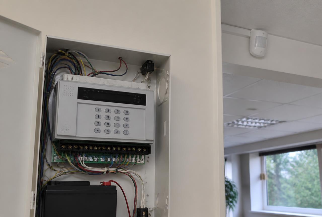 Intrusion alarm control panel with keypad and wiring inside an equipment cupboard, with a ceiling motion detector beyond